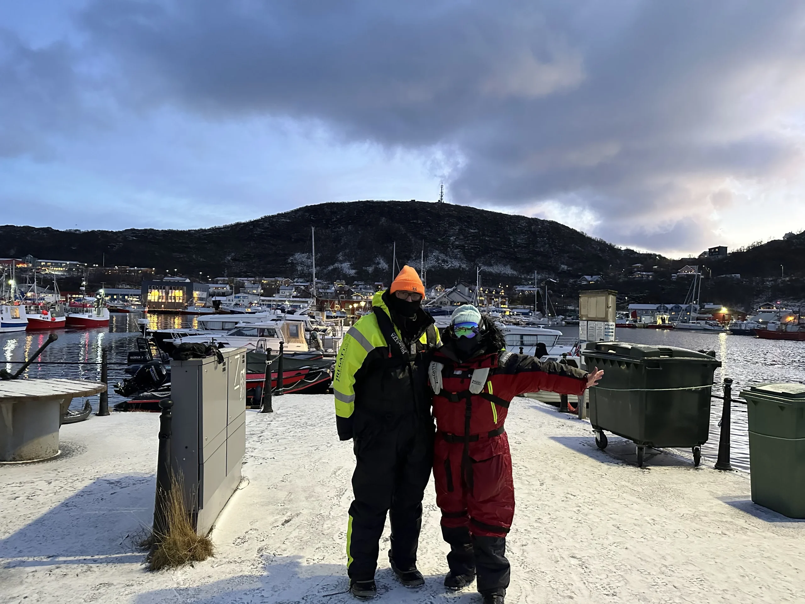 Two people in heavy winter gear stand on a snow-covered dock in Skjervøy, Norway, with a backdrop of a serene harbor, boats, and hills under a dusky sky.