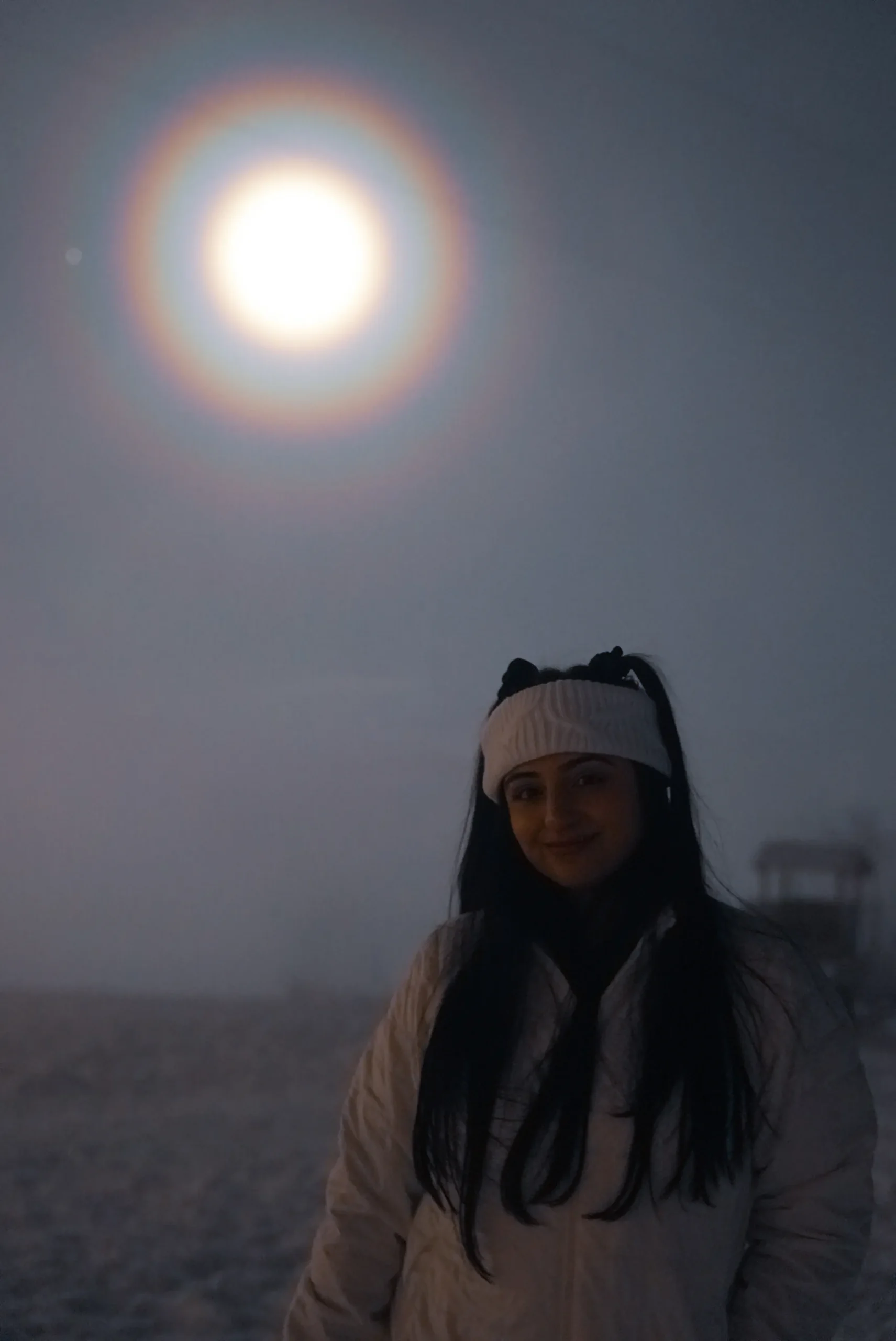 Image of a person standing in the snow at night, facing away from the camera towards a bright moon surrounded by a halo, with anticipation for the northern lights at Fjellheisen.