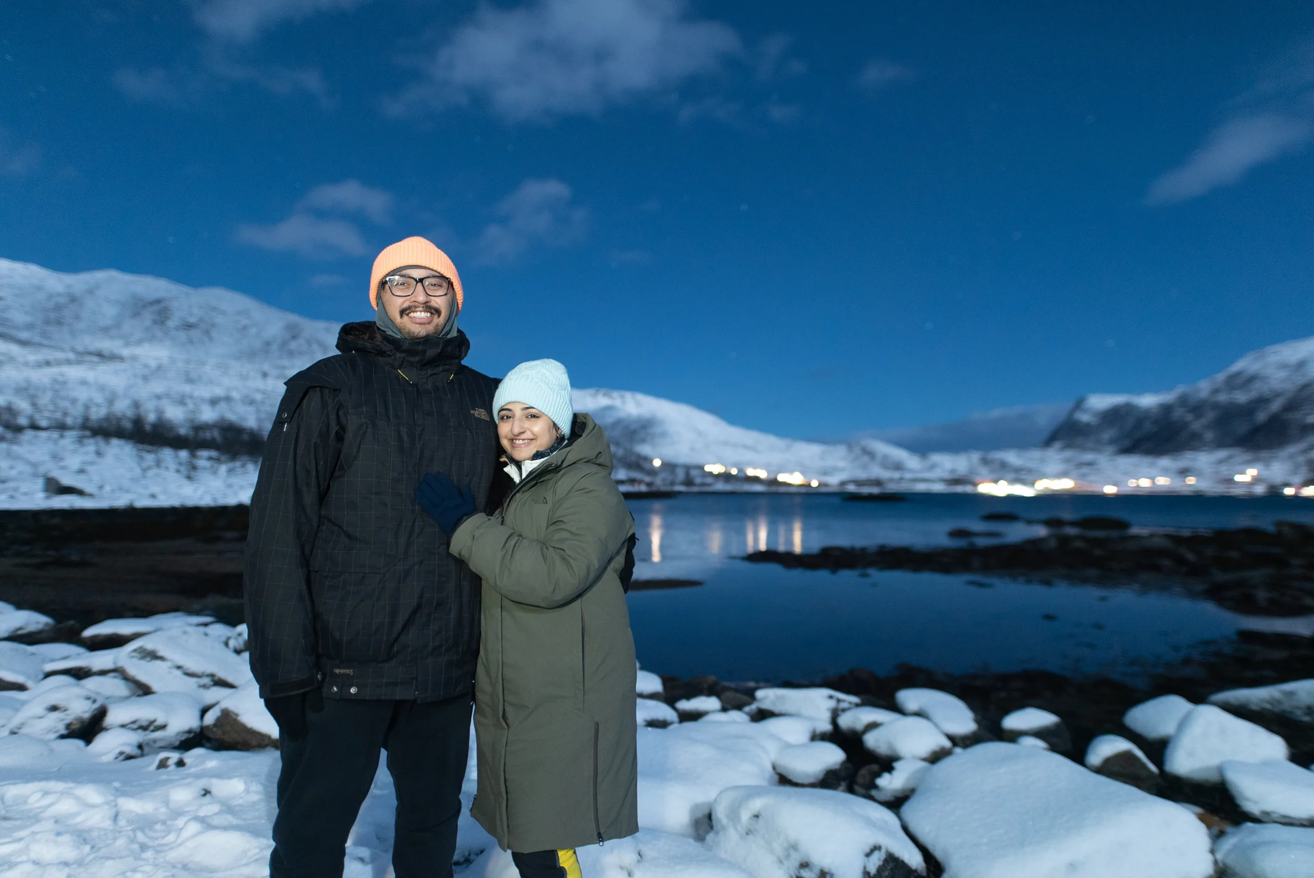 Two people stand with their backs to the camera, gazing at a starry night sky over a snowy landscape in Tromsø, Norway. The Northern Lights are not visible, but anticipation hangs in the air.