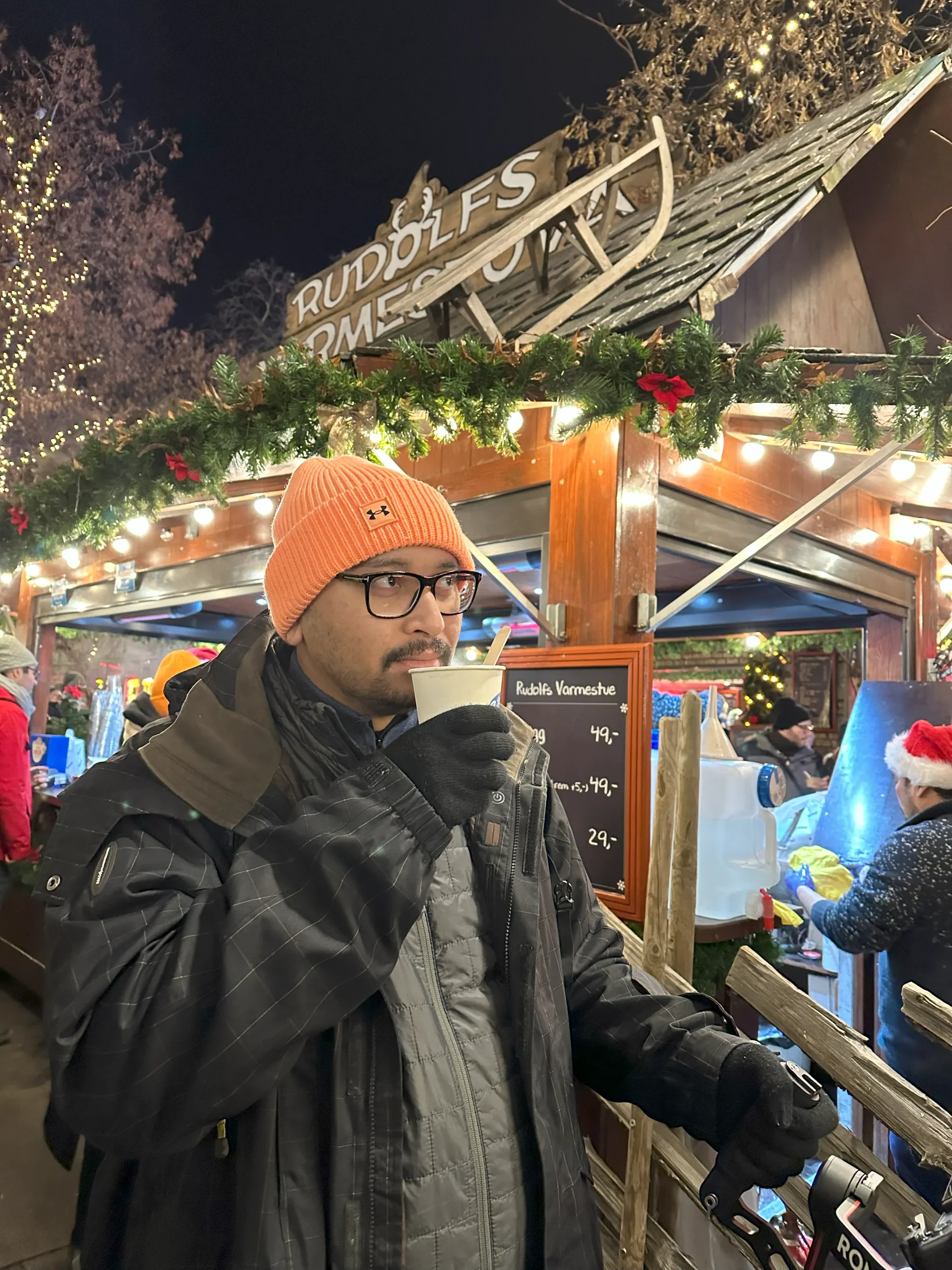 A traveler stands at a festive outdoor market stall adorned with Christmas decorations and lights, holding a cup of mulled wine, with ‘Rudolf’s’ sign overhead and a menu board displaying prices in Norwegian Krone