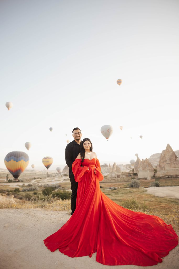 A couple in a red dress and black shirt for photoshoot in Cappadocia, Turkey during sunrise with hot air balloons in the background