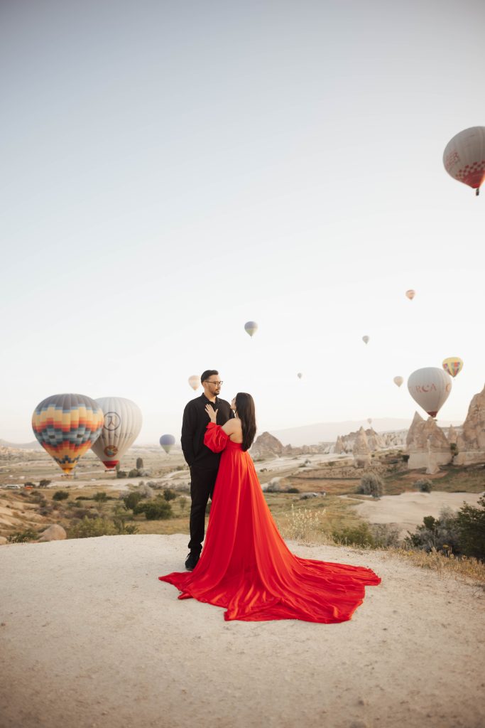A couple in a red dress and black shirt for photoshoot in Cappadocia, Turkey during sunrise with hot air balloons in the background