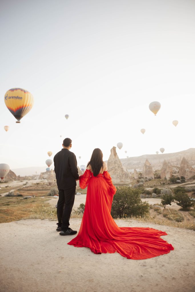 A couple in a red dress and black shirt for photoshoot in Cappadocia, Turkey during sunrise with hot air balloons in the background
