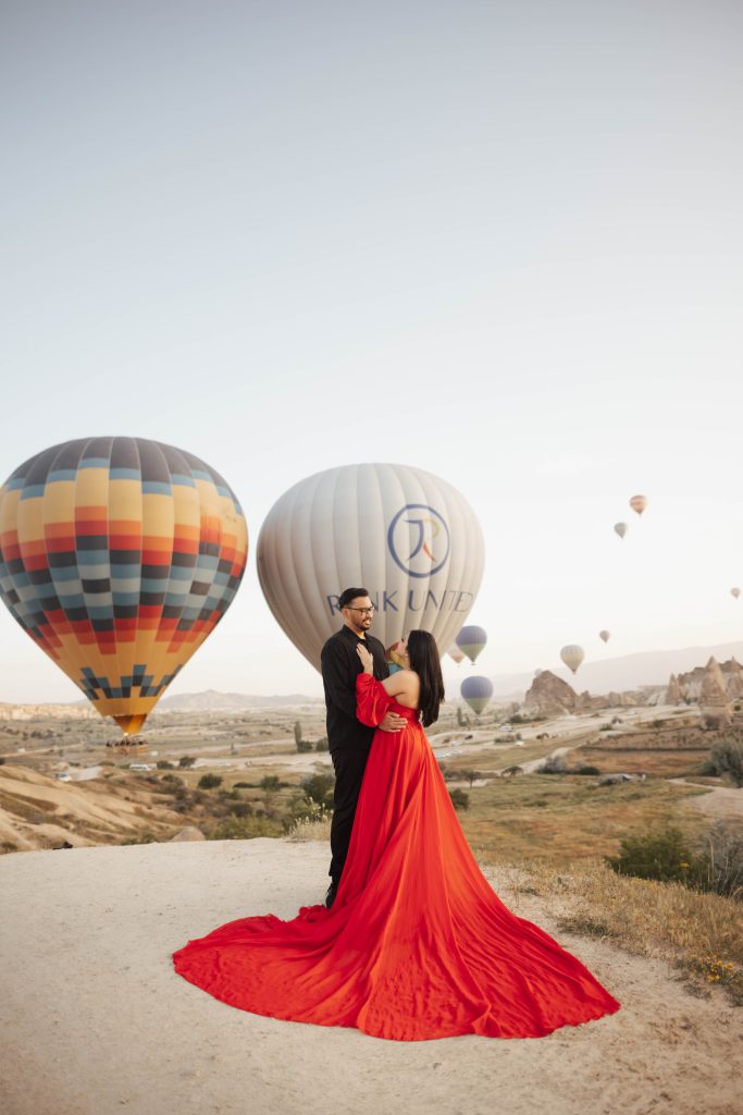 A couple in a red dress and black shirt for photoshoot in Cappadocia, Turkey during sunrise with hot air balloons in the background
