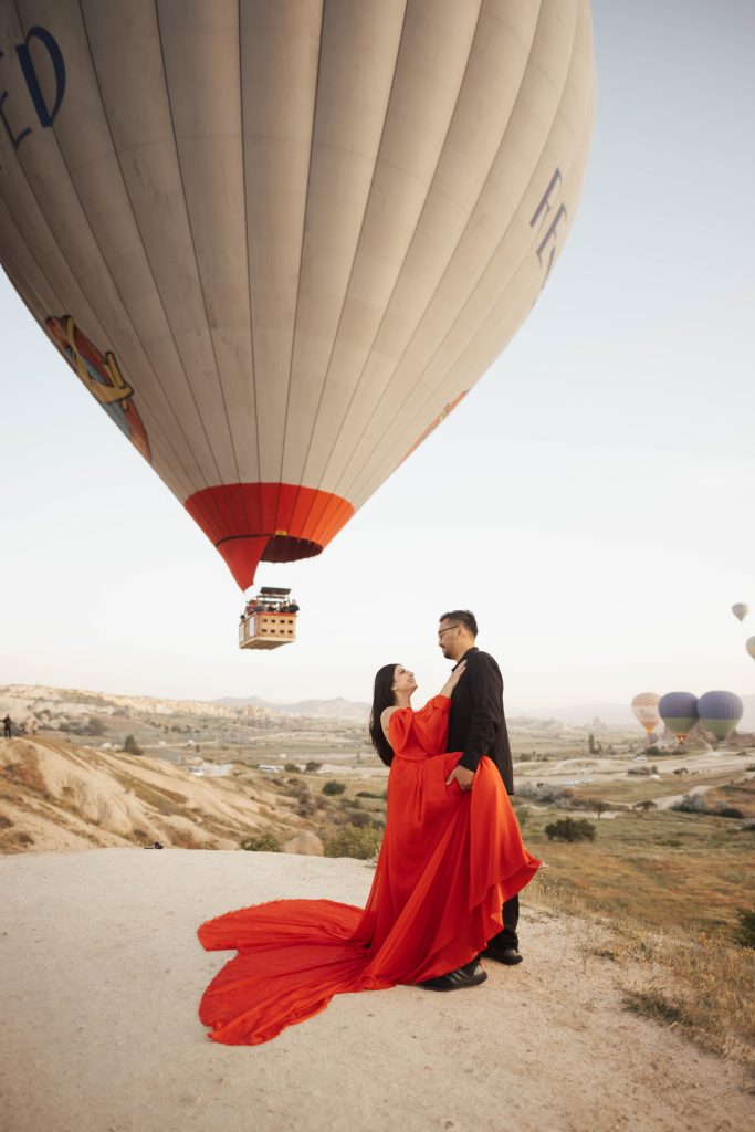 A couple in a red dress and black shirt for photoshoot in Cappadocia, Turkey during sunrise with hot air balloons in the background