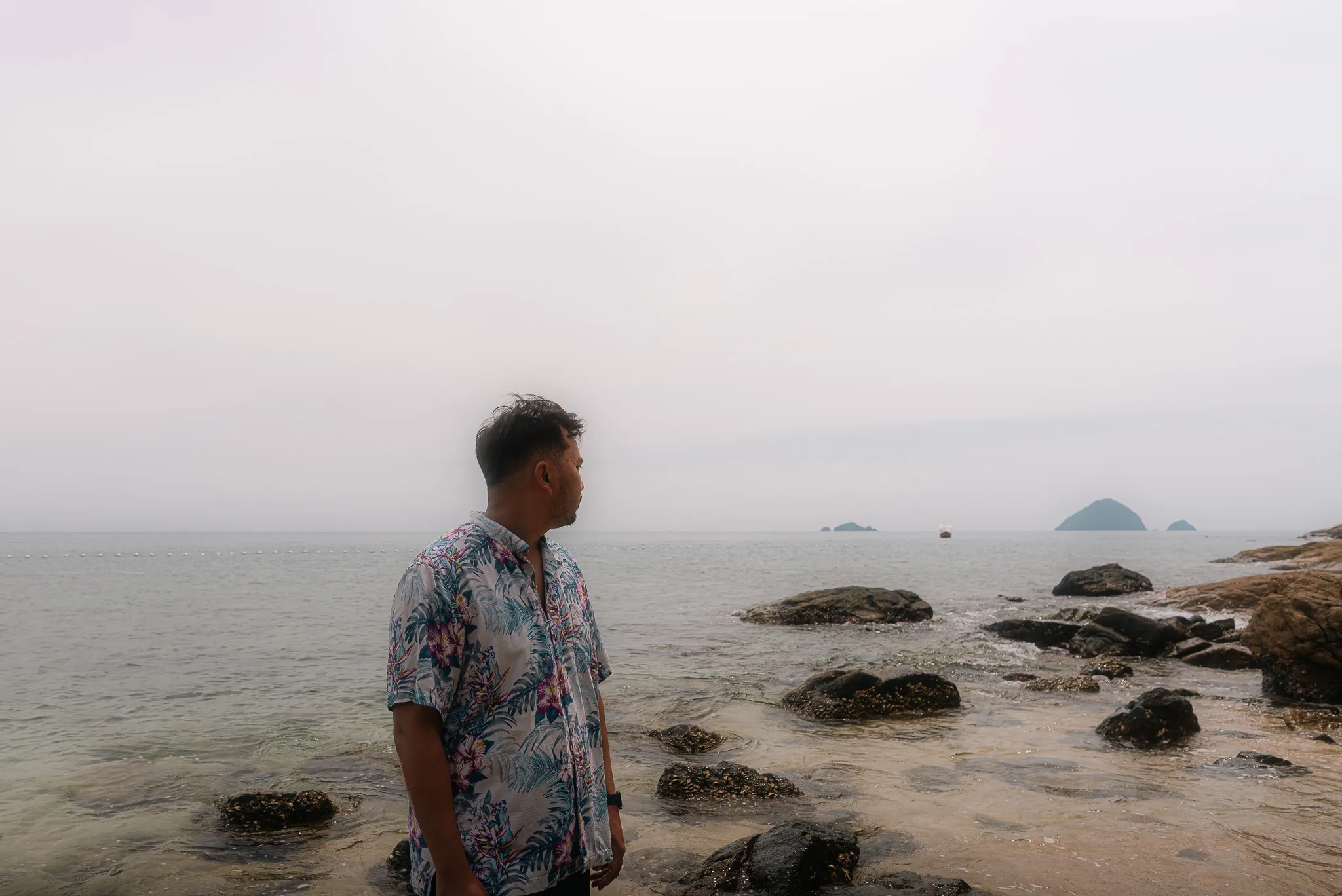 Shariff standing on the rocky shore of Tranquil Beach, looking out at the ocean and distant islands under an overcast sky.