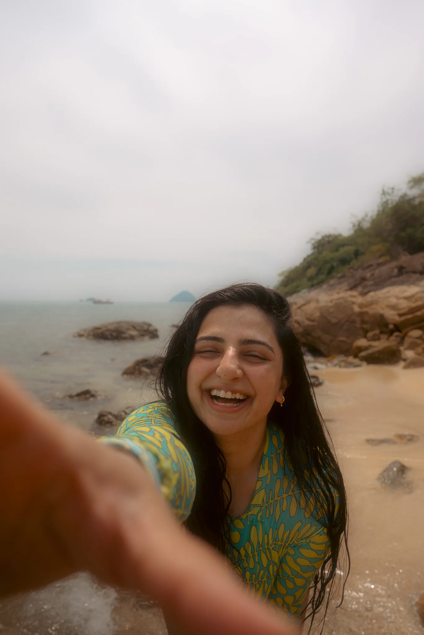 Aroob, a young woman with long dark hair, smiles brightly while taking a selfie on a sandy beach with a hazy ocean in the background.