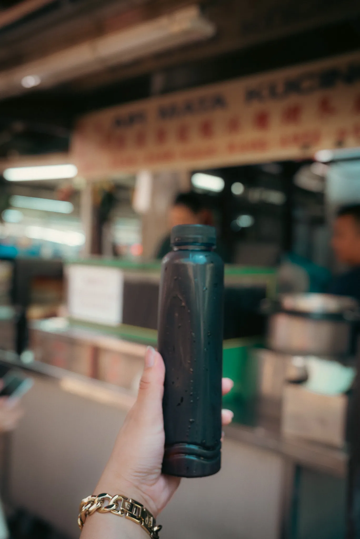 Hand holding a bottle of Air Mata Kucing drink, with the stall in the background, Kuala Lumpur
